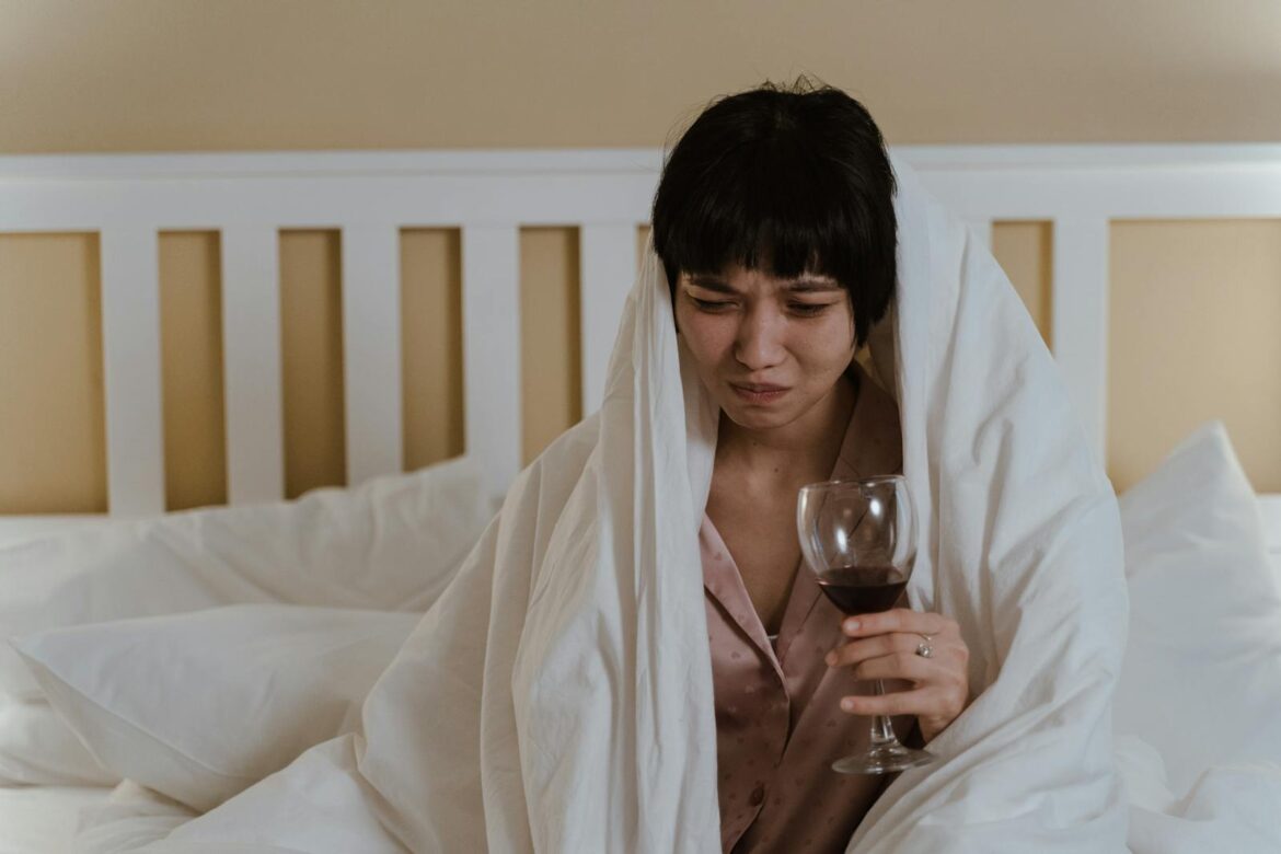 A woman wrapped in a blanket holding a wine glass, showing a look of discomfort.