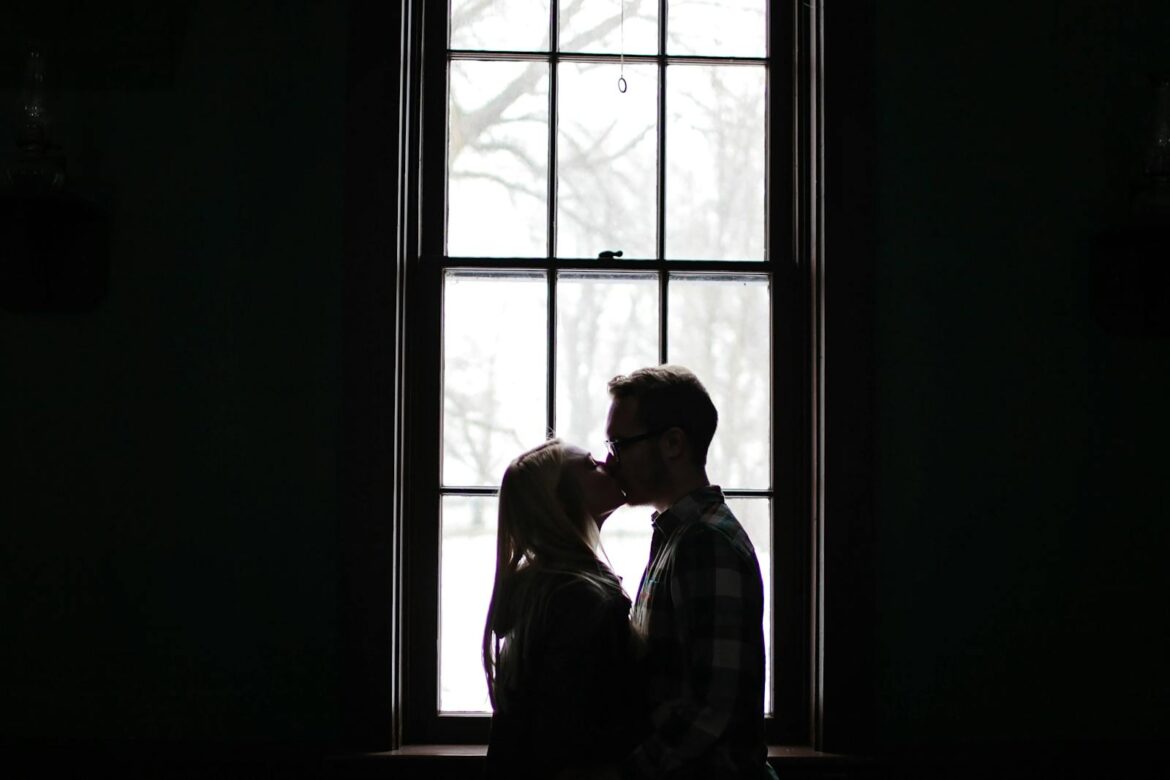 A romantic silhouette of a couple sharing a kiss in front of a large, backlit window.
